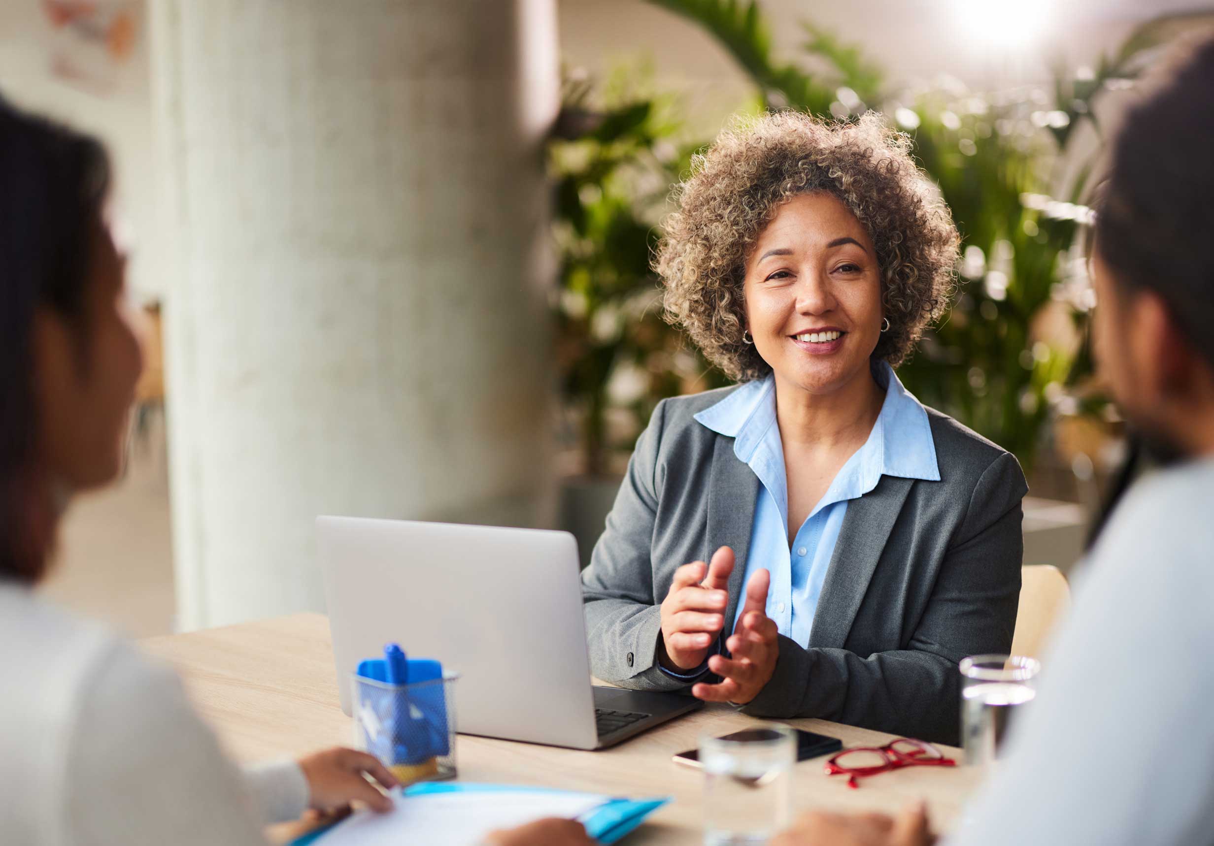 A Novaris Offshore Bank financial advisor with short curly hair is smiling and gesturing while seated at a table with her computer. She is meeting with a client to discuss their financial needs, symbolizing the expert and personalized service we offer.